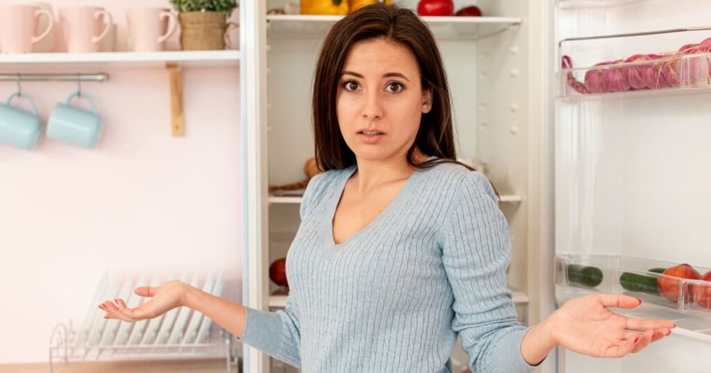Woman staring blankly into an open refrigerator feeling stressed about what to cook for dinner.