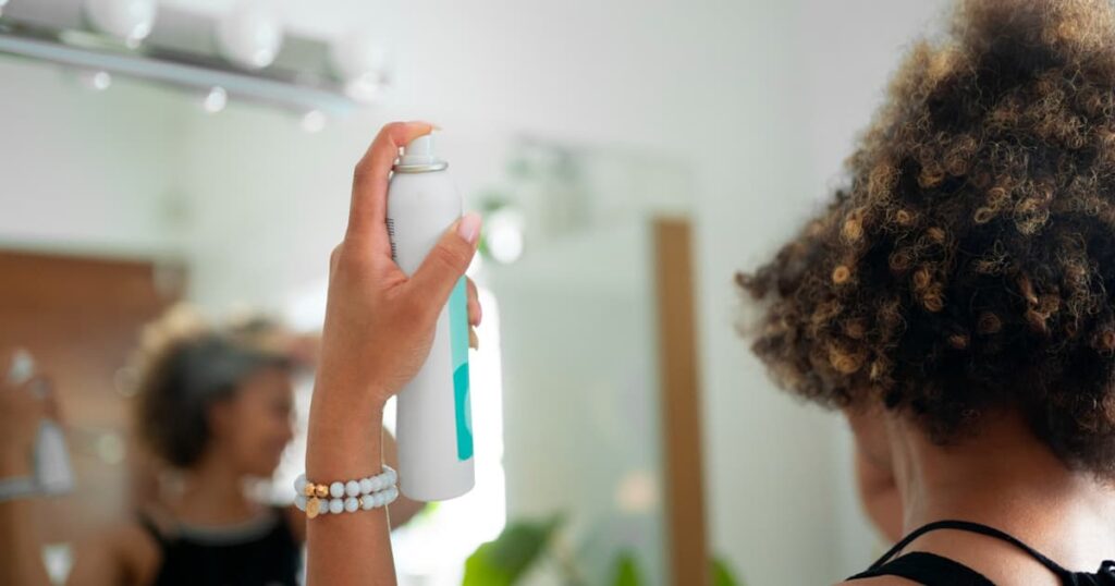 A can of dry shampoo being sprayed onto hair roots to refresh them without water.