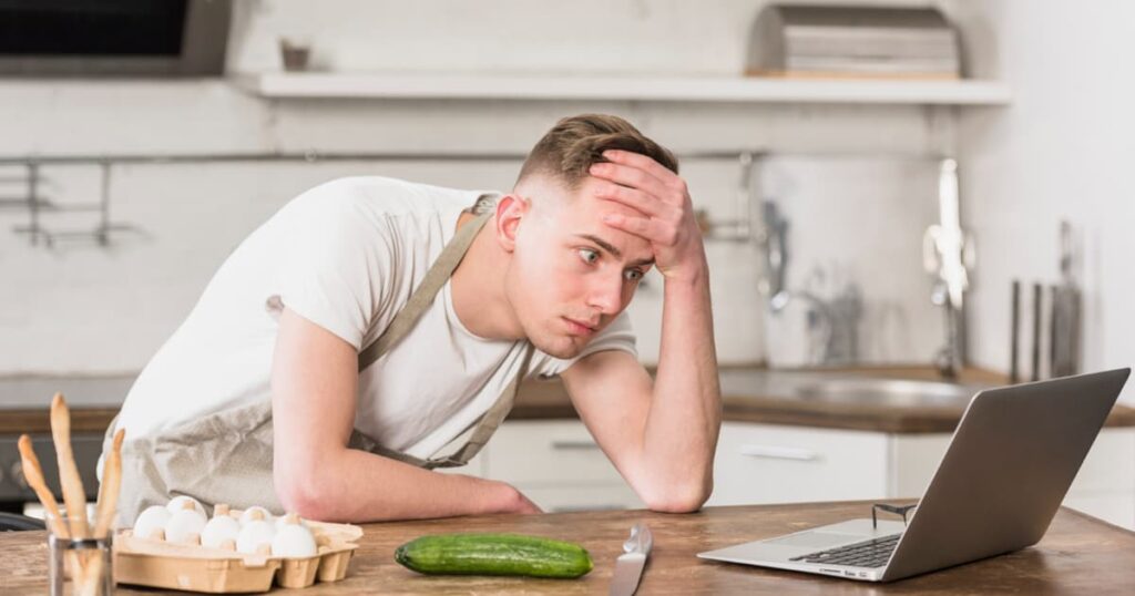 A person trying to type on a laptop while cooking and talking on the phone, looking stressed.