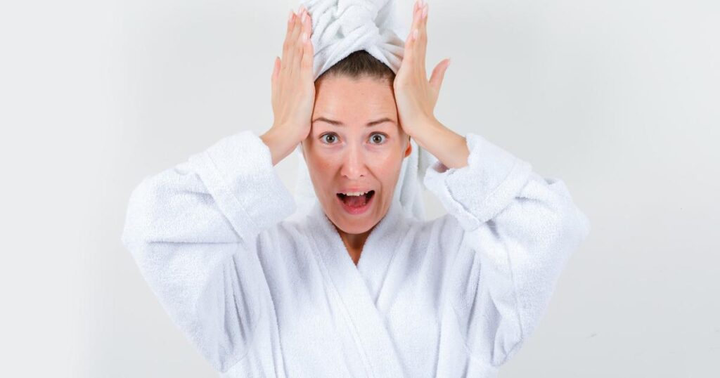 A person wrapped in a towel shivering and looking angry at a shower head.