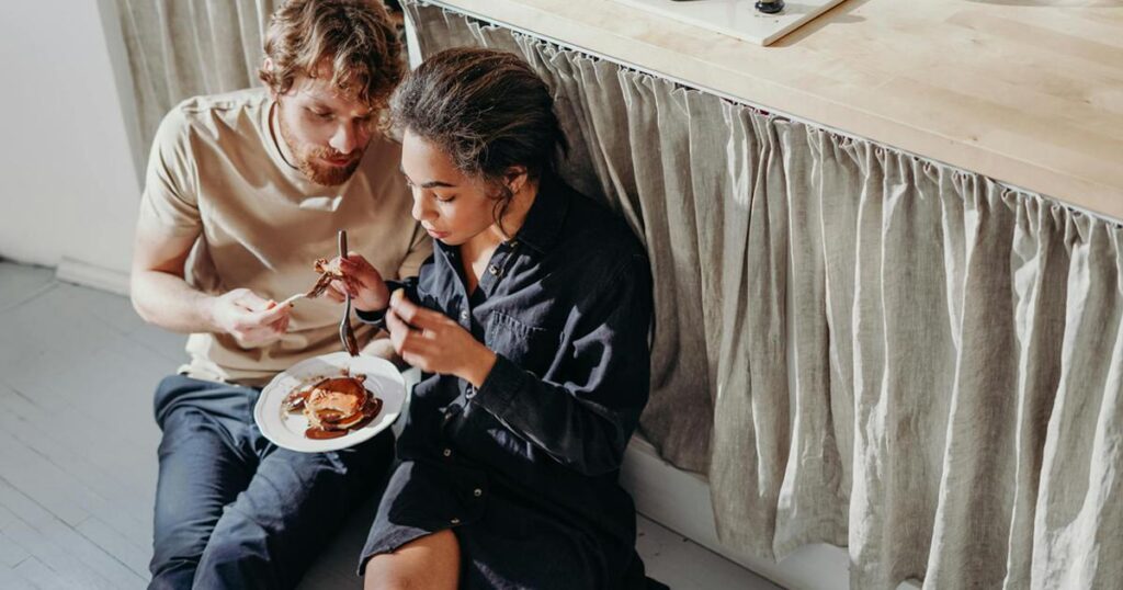 A couple eating takeout food on the floor in sweatpants, relaxing without pressure.