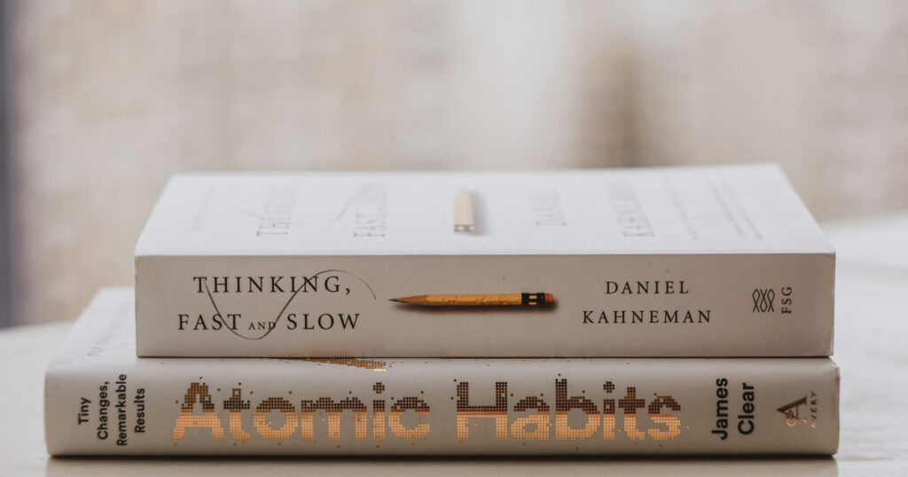 A cozy aesthetic stack of the best books for personal growth, including Atomic Habits and The Mountain Is You, sitting on a wooden table next to a cup of coffee.